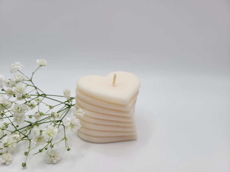 May include: A white heart-shaped candle with multiple layers. The candle is sitting on a white surface with a sprig of white flowers.