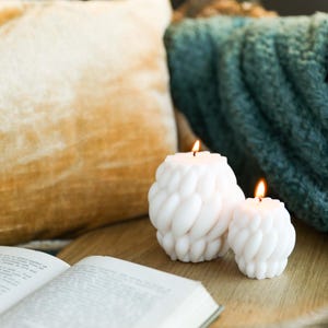 May include: Two white, textured candles with a flame burning on each. The candles are on a wooden surface with a book and a throw blanket in the background.