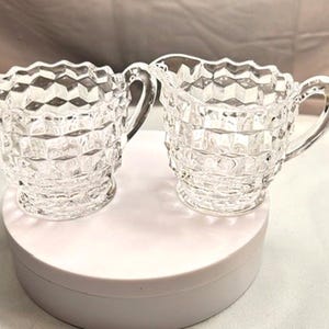 May include: Two clear glass cream and sugar set. Each piece has a diamond pattern and a scalloped rim. The sugar bowl has two handles, and the creamer has a handle and a spout. The set is displayed on a white surface.