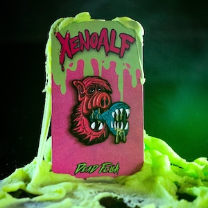 May include: A pink and green enamel pin featuring a cartoon pig wearing a blue and green helmet. The pin says "Xenoalf" and "Dead Fella" on it.