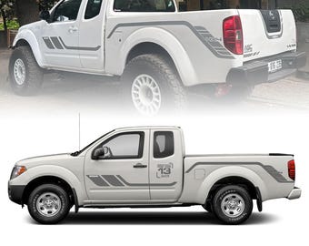 May include: A white Nissan Frontier pickup truck with black and gray graphic decals on the sides. The truck has a black bed liner and a black step bar. The truck is parked on a paved surface.