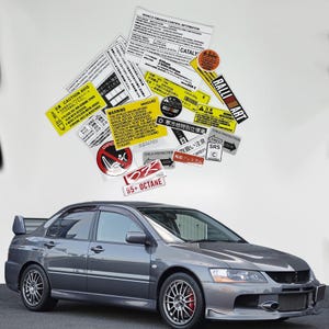 May include: A gray sports car with a rear spoiler and red brake calipers. Above the car are various stickers with text, including "CAUTION AVIS", "RALLI ART", and "95+ OCTANE". The stickers are in multiple languages.
