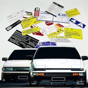 May include: Two white Japanese cars with various warning stickers and labels on the windshield. The stickers are in Japanese and English and include information about tire pressure, engine capacity, and safety warnings. The cars are both facing forward and have black bumpers.