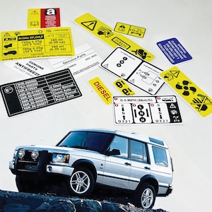 Puede incluir: Un Land Rover Discovery SUV plateado con detalles negros y un parachoques delantero negro. El vehículo está estacionado en una superficie rocosa. Varios adhesivos de advertencia amarillos y negros son visibles en el fondo blanco. Los adhesivos contienen texto y símbolos relacionados con el mantenimiento y la seguridad del vehículo.