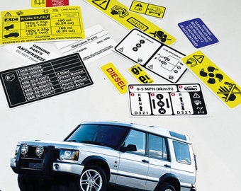 Peut inclure: Un Land Rover Discovery SUV argenté avec des garnitures noires et un pare-chocs avant noir. Le véhicule est garé sur une surface rocheuse. Plusieurs autocollants d'avertissement jaunes et noirs sont visibles sur le fond blanc. Les autocollants contiennent du texte et des symboles liés à l'entretien et à la sécurité du véhicule.