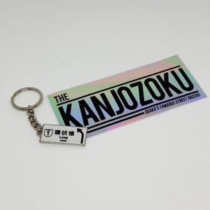 Könnte beinhalten: Ein rechteckiger Schlüsselanhänger mit einem weißen Schild, einem schwarzen Pfeil, der Zahl 1 und japanischem Text, befestigt an einem Metallring. Daneben liegt ein rechteckiger Aufkleber mit holografischem Finish und dem Text THE KANJOZOKU OSAKA'S FAMAOUS STREET RACERS.