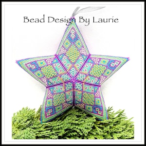 May include: A colourful beaded star ornament with a geometric pattern in shades of purple, green, yellow, and blue. The star is hanging from a white string and is resting on a bed of green foliage. The text "Bead Design By Laurie" is visible at the top of the image.