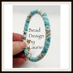 May include: A beaded bracelet with a magnetic clasp. The bracelet is primarily turquoise with accents of silver and brown beads. The text "Bead Design By Laurie" is visible on the bracelet.