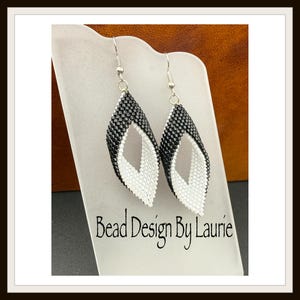 May include: Handmade earrings featuring a black and white beaded pattern. The earrings are teardrop-shaped, with a white central void. The text "Bead Design By Laurie" is displayed below the earrings.