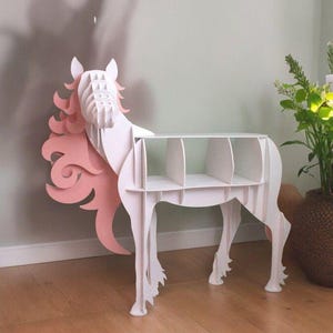 Puede incluir: Estantería de madera blanca en forma de unicornio con melena y cola rosas. La estantería tiene cuatro estantes.