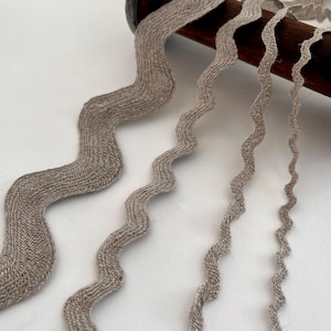 May include: A close-up shot of several wavy ribbons in varying widths. The ribbons are a light brown colour and have a textured, woven appearance. They are arranged in a flowing pattern against a white background. The ribbons are likely for crafts or decoration.