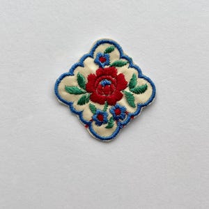 Puede incluir: Parche bordado con diseño floral. El parche presenta un fondo color crema con una rosa roja en el centro, rodeada de hojas verdes y detalles azules. El parche está delineado con un borde azul.