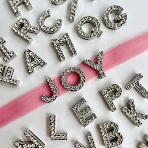 May include: Silver letter charms with rhinestones, spelling out the word "JOY". The charms are scattered on a pink ribbon and a white surface.