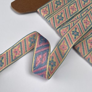 May include: Woven ribbon featuring a repeating pattern of pink and blue floral diamond shapes on a yellow background. The ribbon is folded and curved, showcasing the texture and detail of the design. A small piece of brown material is visible in the background.