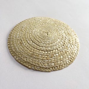 May include: A round, woven straw placemat. The placemat is a light beige color and has a spiral pattern in the center. The straw is tightly woven, creating a textured surface. This item is suitable for home decor.