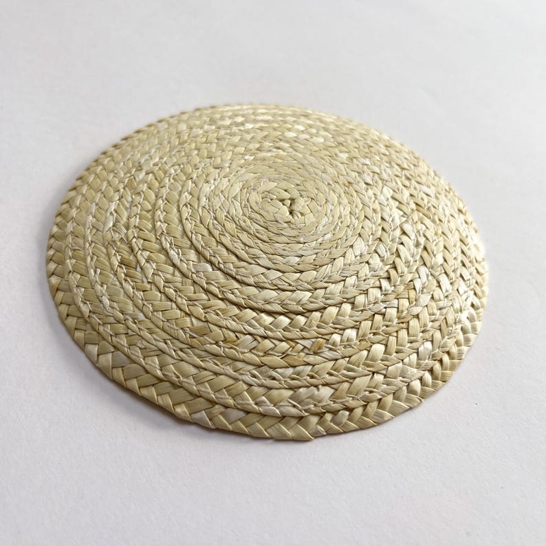May include: A round, woven straw placemat. The placemat is a light beige color and has a spiral pattern in the center. The straw is tightly woven, creating a textured surface. This item is suitable for home decor.