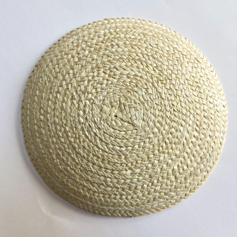 May include: A round, woven straw hat with a shallow crown. The hat is made of natural-colored straw and has a tightly woven pattern. The brim is slightly curved, and the hat appears to be handmade. The hat is likely for sun protection or fashion.
