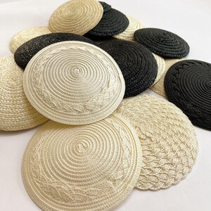 May include: A collection of woven kippahs in cream and black. The kippahs have a circular design with a spiral pattern and some feature a decorative leaf pattern. The hats are made of a woven material.