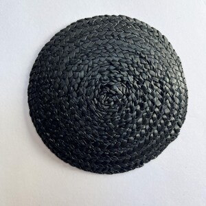 May include: A round, black woven coaster. The coaster is made of tightly woven, dark material, creating a textured surface. The weave pattern spirals inward from the outer edge to the center.
