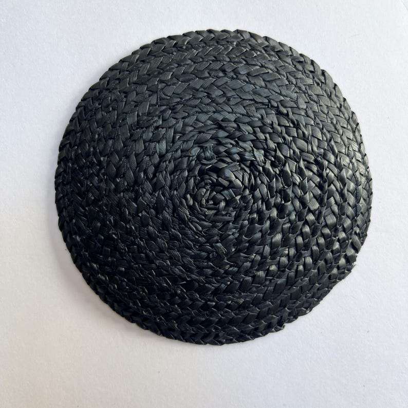 May include: A round, black woven coaster. The coaster is made of tightly woven, dark material, creating a textured surface. The weave pattern spirals inward from the outer edge to the center.