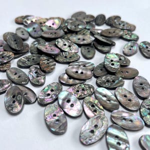 Può includere: Un insieme di bottoni ovali con due fori ciascuno. I bottoni hanno una finitura perlescente e iridescente con sfumature di grigio, verde e viola. I bottoni sono sparsi su una superficie bianca.