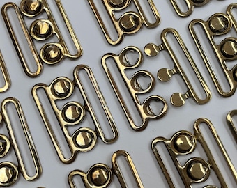May include: A collection of brass-toned metal hook and eye fasteners arranged in a grid pattern. Each fastener consists of a rectangular frame with three circular loops on one side and a corresponding set of three hooks on the other side.