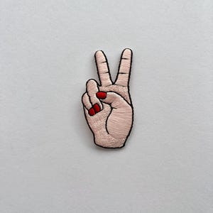 Puede incluir: Parche bordado que representa una mano haciendo el signo de la paz. La mano es de tono claro, con uñas rojas y contorno negro. El parche es para ropa o accesorios.
