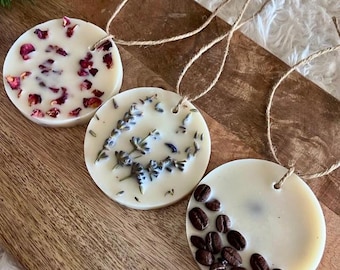 May include: Three circular wax melts in cream, each decorated with natural elements. One features rose petals, another lavender, and the third coffee beans. All have a jute string for hanging.