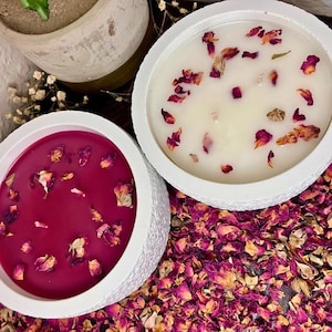 May include: Two white, textured, round candles, one with a deep red wax and the other with a creamy white wax. Both are decorated with dried rose petals. The candles sit on a bed of dried rose petals.