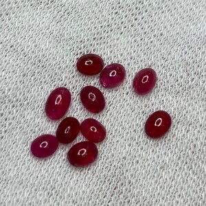 May include: A collection of ten oval-shaped, red gemstones on a white fabric background.