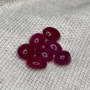 Op de afbeelding: Zeven kleine, ovale, dieprode edelstenen op een witte stoffen achtergrond.