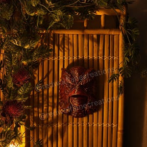 May include: A dark red, Creature from the Black Lagoon mask is mounted on a bamboo wall. The mask is detailed with textured features. The wall is decorated with tropical leaves and red flowers.