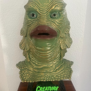 May include: A green and gold Creature from the Black Lagoon mask. The mask has a textured surface with detailed features, including large eyes and a prominent mouth. The base of the mask rests on a wooden box with the text "CREATURE FROM THE BLACK LAGOON".