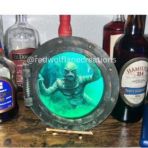 May include: A round, dark metal framed wall art piece featuring a Creature from the Black Lagoon design. The creature is green and the background is a teal color. The text "@redwolflanecreations" is visible on the art piece.