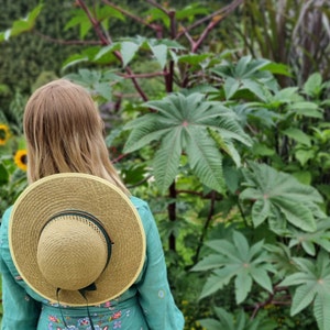 May include: A woman wearing a teal dress with a floral pattern and a wide-brimmed straw hat with a green band stands in a garden with lush green foliage.