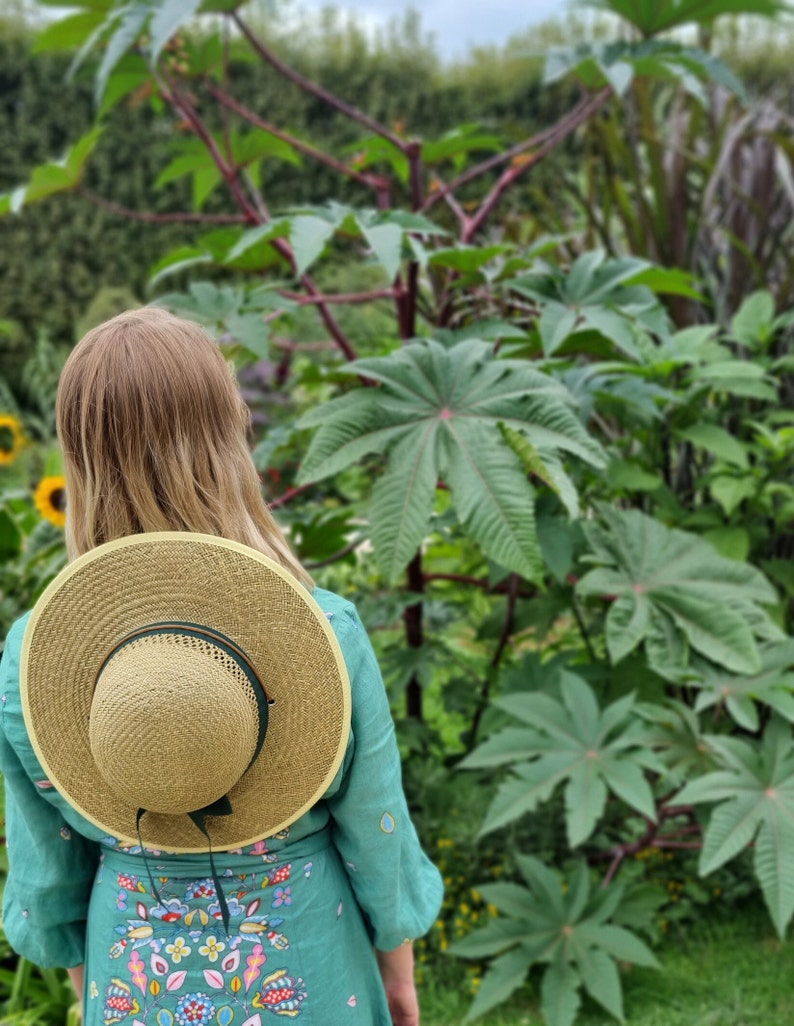 May include: A woman wearing a teal dress with a floral pattern and a wide-brimmed straw hat with a green band stands in a garden with lush green foliage.