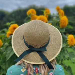 May include: A wide-brimmed straw hat with a green ribbon and bow. The hat is worn by a person standing in a field of yellow flowers.