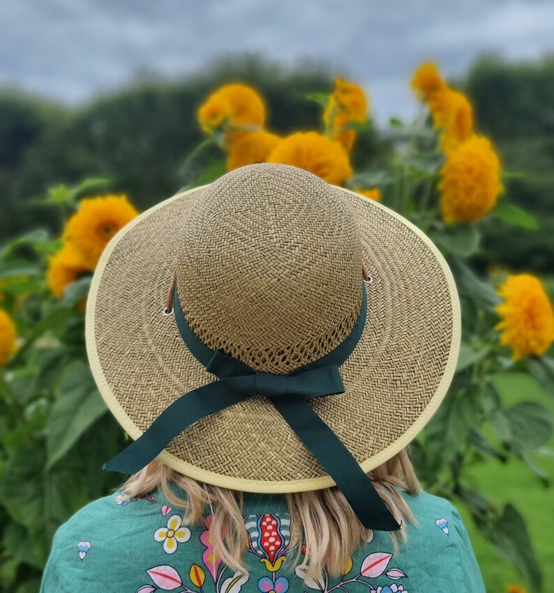 May include: A wide-brimmed straw hat with a green ribbon and bow. The hat is worn by a person standing in a field of yellow flowers.