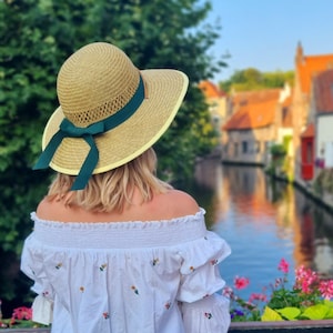 May include: A woman wearing a white off-the-shoulder blouse with floral embroidery, a wide-brimmed straw hat with a green ribbon, and blonde hair. She is standing on a bridge overlooking a canal with colorful buildings in the background.