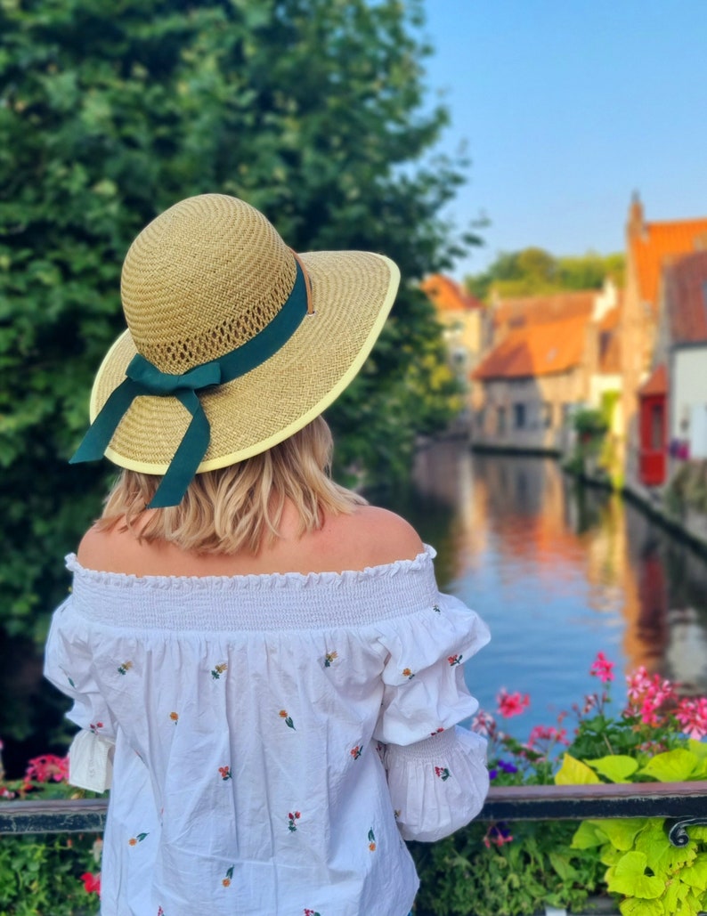 May include: A woman wearing a white off-the-shoulder blouse with floral embroidery, a wide-brimmed straw hat with a green ribbon, and blonde hair. She is standing on a bridge overlooking a canal with colorful buildings in the background.