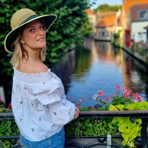May include: A woman wearing a straw hat with a green band, a white off-the-shoulder blouse with floral embroidery, and blue jeans, stands on a bridge overlooking a canal in a European city.