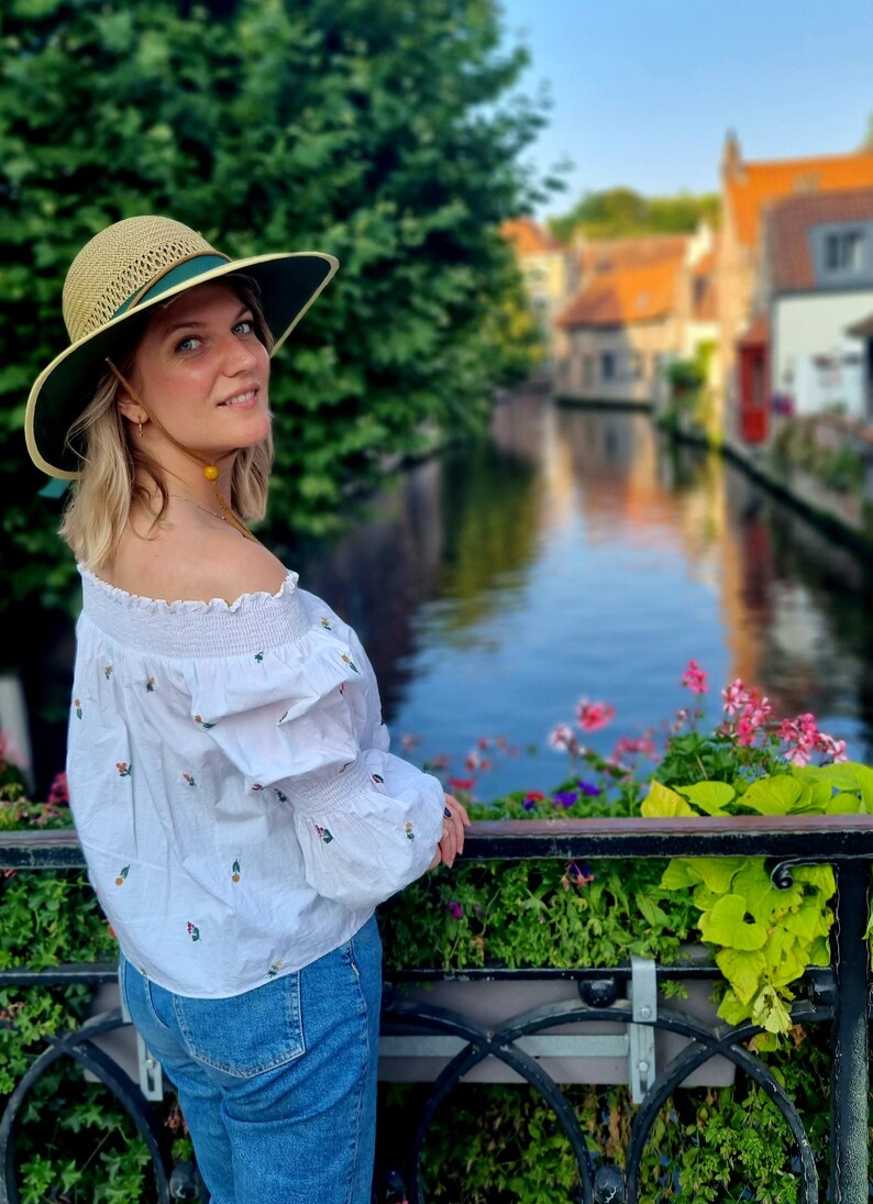 May include: A woman wearing a straw hat with a green band, a white off-the-shoulder blouse with floral embroidery, and blue jeans, stands on a bridge overlooking a canal in a European city.