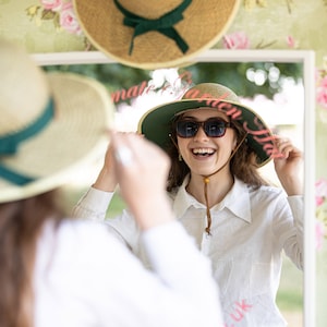 May include: A woman tries on a wide-brimmed straw hat with a green band and pink lettering that says "Make it a Garden Hat UK". She is smiling and looking at herself in a mirror.