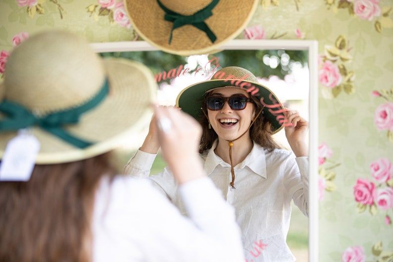 May include: A woman tries on a wide-brimmed straw hat with a green band and pink lettering that says "Make it a Garden Hat UK". She is smiling and looking at herself in a mirror.