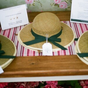 May include: Three straw hats with green ribbon bows and white tags. The hats are displayed on a wooden table with a striped tablecloth. The text on the tag reads "The Royal Horticultural Society 5 Star Trademark Award".