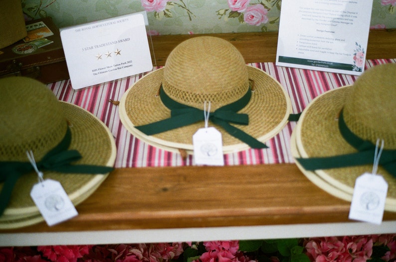 May include: Three straw hats with green ribbon bows and white tags. The hats are displayed on a wooden table with a striped tablecloth. The text on the tag reads "The Royal Horticultural Society 5 Star Trademark Award".