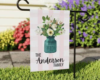 May include: A garden flag featuring a watercolor illustration of white flowers in a teal vase. The background has a pink and white striped pattern. The text "The Anderson Family" is displayed in black script.