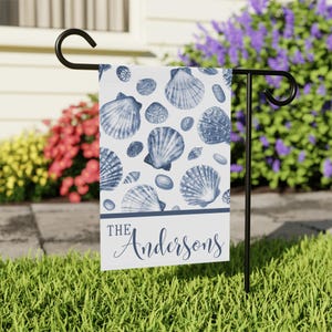 May include: A white garden flag featuring a pattern of blue seashells. The flag has a horizontal blue band with the name "The Andersons" in a script font. The flag is displayed on a black metal stand in a grassy yard.