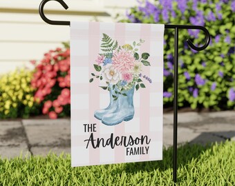 May include: A garden flag featuring a pair of light blue rain boots filled with a bouquet of pink, white, and blue flowers. The flag has a pink and white striped background and reads "The Anderson Family" in black script.