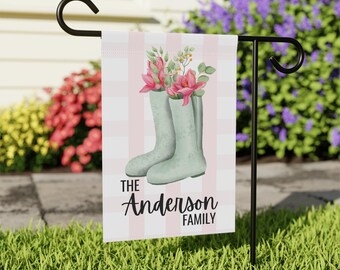 May include: A garden flag featuring a pair of light green rain boots filled with pink flowers. The background has a pink and white striped pattern. The text "The Anderson Family" is displayed in black script.